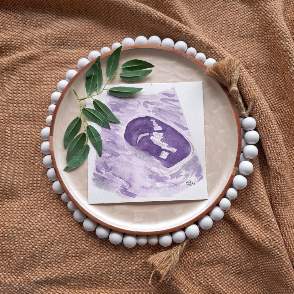 Round ceramic plate with decorative beads, a card with a purple watercolour ultrasound, and green leaves on a brown fabric background.