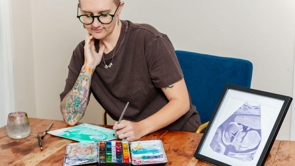 A tattooed person wearing glasses sits at a wooden table, holding a paintbrush over an open watercolor set. A framed ultrasound-style print is on the table beside them, with a blue chair and light wall in the background.