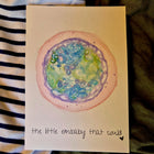 Card with a watercolor illustration of an embryo and text 'the little embaby that could' on a striped fabric background.