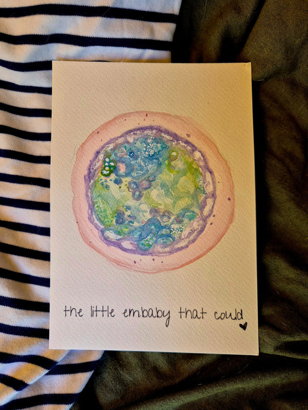 Card with a watercolor illustration of an embryo and text 'the little embaby that could' on a striped fabric background.