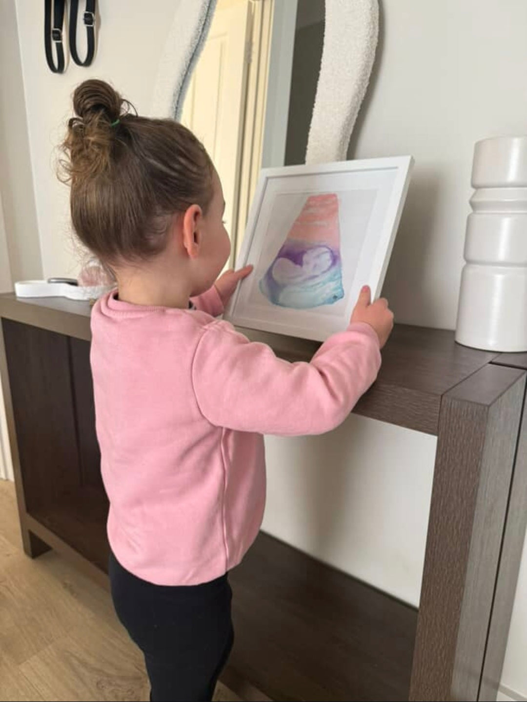 Child in a pink jacket holding a framed watercolour sonogram on a wooden surface.