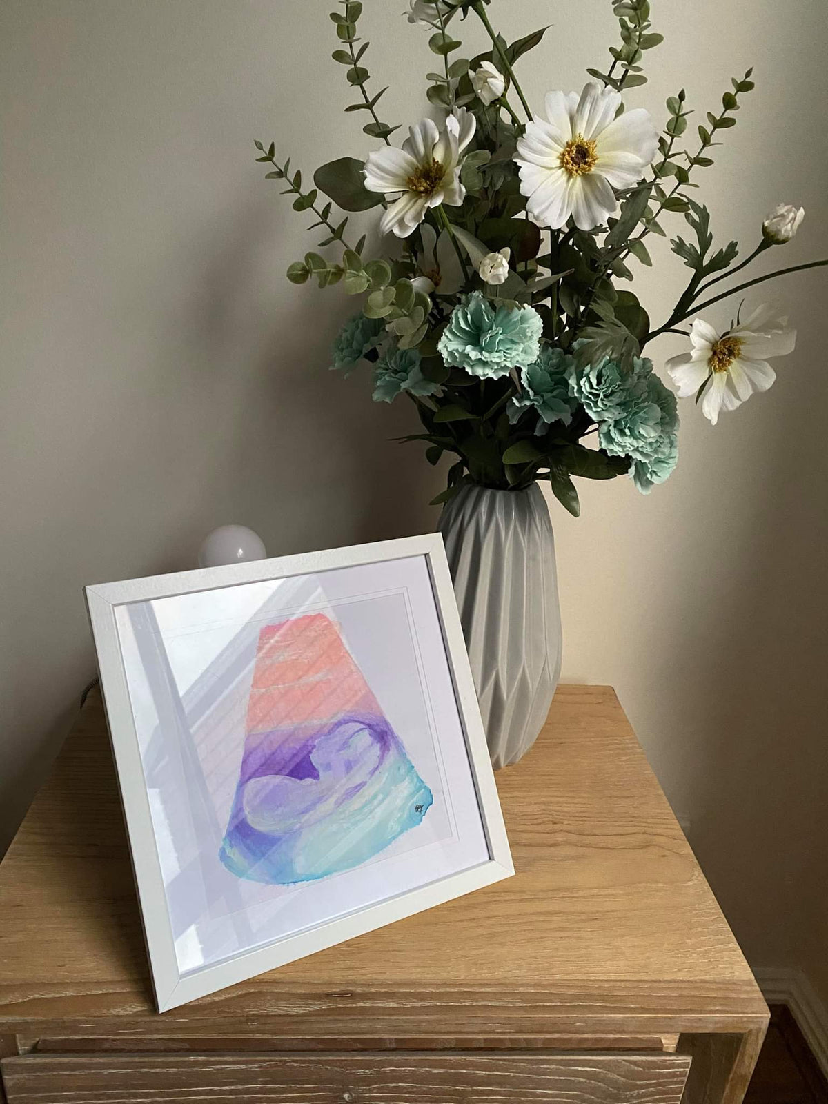 Framed pink purple and blue 12 week sonogram painting on a wooden surface with a vase of flowers in the background