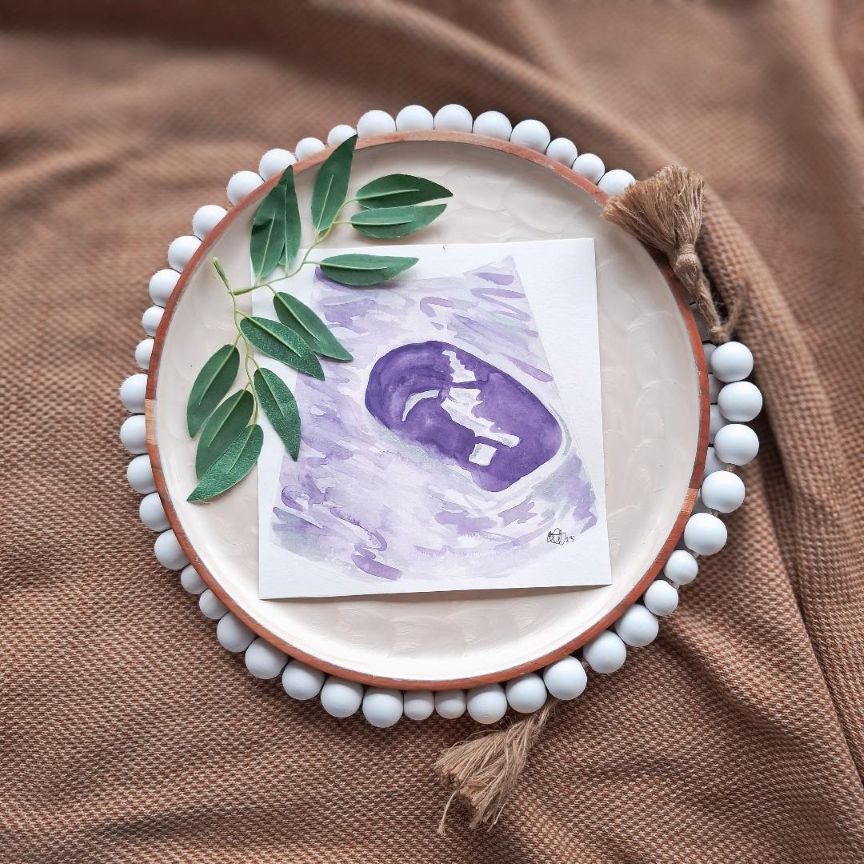 Round ceramic plate with white beads around the edge, featuring a purple early pregnancy ultrasound watercolour painting and green leaves on a brown fabric background.