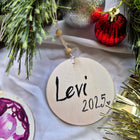 Round wooden ornament with 'Levi 2025' text, surrounded by Christmas decorations.