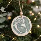 A round wooden Christmas ornament with a hand-painted green and white ultrasound, hanging from a twine string with a wooden bead, set against a blurred Christmas tree background with warm string lights.