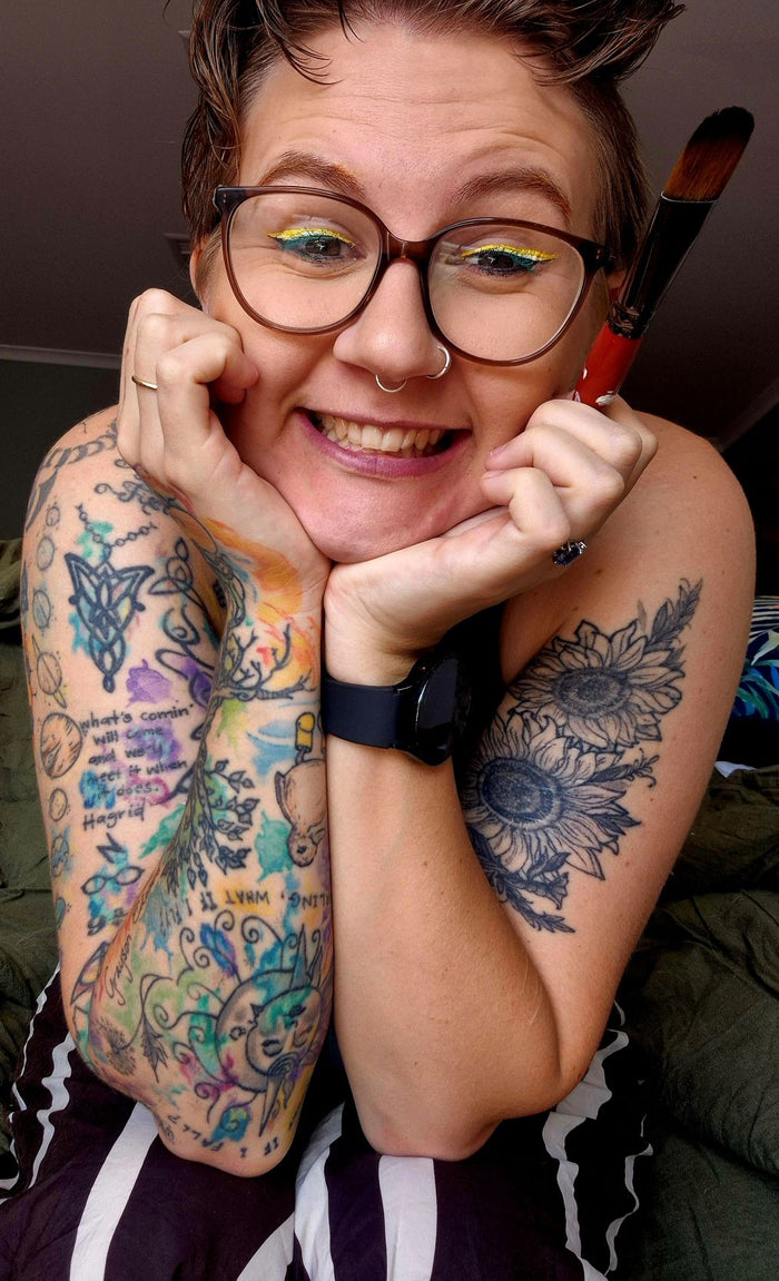 Person with tattoos on arms wearing glasses and a striped shirt, with a brush in the background.