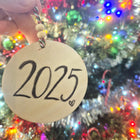 Round ornament with '2025' text held in front of a Christmas tree with lights.