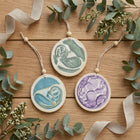 Three round ceramic Christmas ornaments with wooden beads and jute hanging strings, featuring hand-painted baby motifs in green, blue, and purple, arranged on a wooden surface with eucalyptus leaves and ribbon.