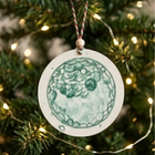 A round ceramic Christmas ornament featuring a green watercolor embryo design, hanging from a red-and-white striped string on a decorated Christmas tree with warm lights.