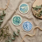 Three round wooden hanging ornaments with watercolor-style blue and green designs, arranged on a beige fabric surface with ribbon and eucalyptus sprigs.