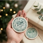 A hand holds a round wooden ornament with a green watercolor illustration of an embryo, with another matching ornament resting on wrapping paper beside a wrapped gift box and a blurred Christmas tree in the background.