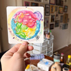 Hand holding a watercolour embryo painting in a room with various items on a desk.