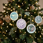 Three round ceramic Christmas ornaments with hand-painted designs hang on a decorated Christmas tree with warm string lights. One ornament is light blue with a green and white design, one is white with a lavender illustration, and one is white with a red and green holiday design.