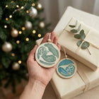 A hand holds two round wooden ornaments with hand-painted baby ultrasounds, one in green and one in blue, in front of wrapped Christmas gifts and a decorated tree.