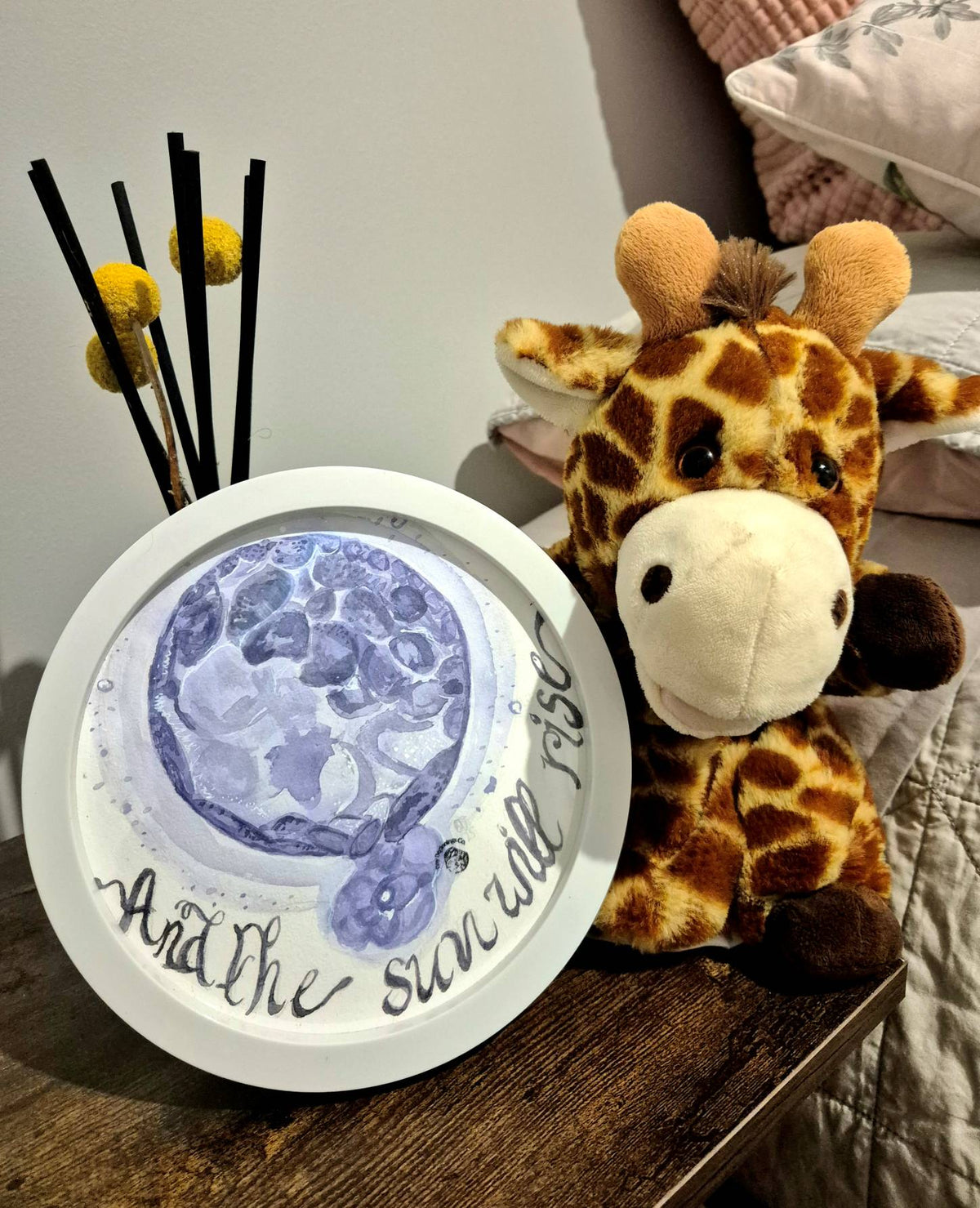 watercolour embryo painting with text 'and the sun will rise', on bedside table with stuffed giraffe toy